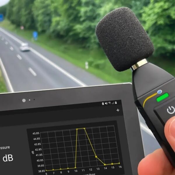 Person holding a sound level meter with a microphone attached, measuring noise levels on a tablet showing a decibel chart. The background depicts a highway with vehicles and greenery.