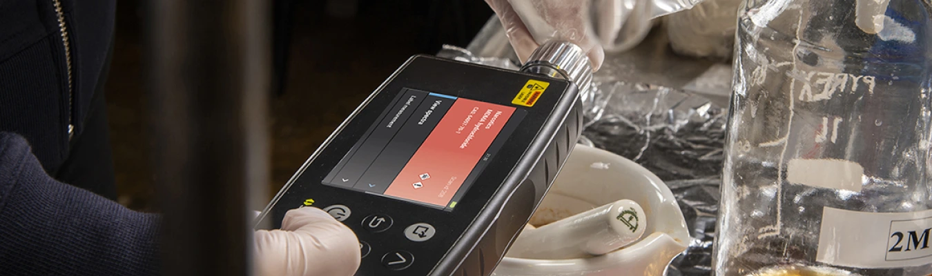 A person wearing gloves uses a portable analysis device with a display screen showing measurements. The device is positioned near a mortar and pestle, and a large container labeled "2M". The setting appears to be a laboratory or a workspace with scientific equipment.